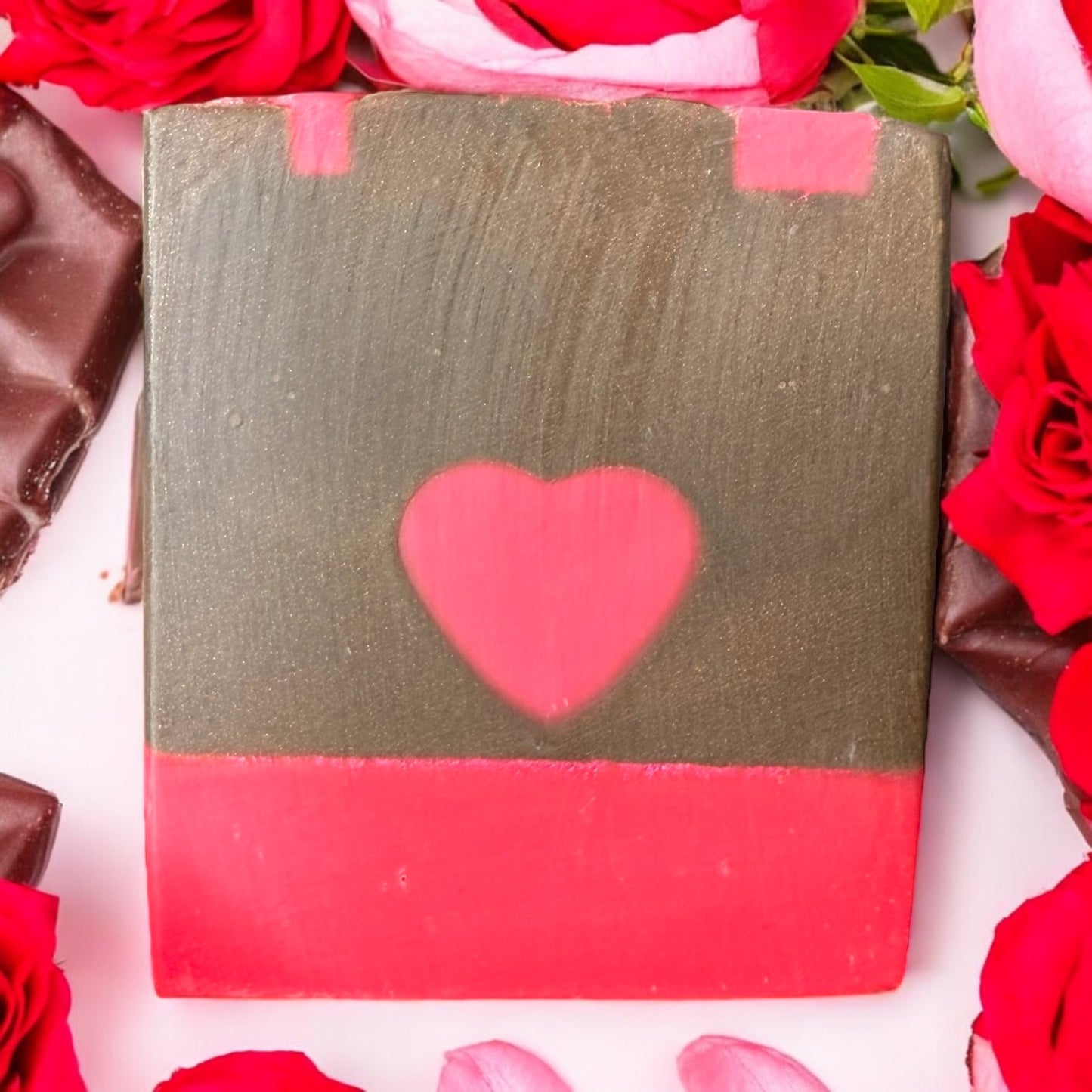 A bar of soap with a chocolate brown color and a red heart design on top and in the middle, presented alongside red roses.