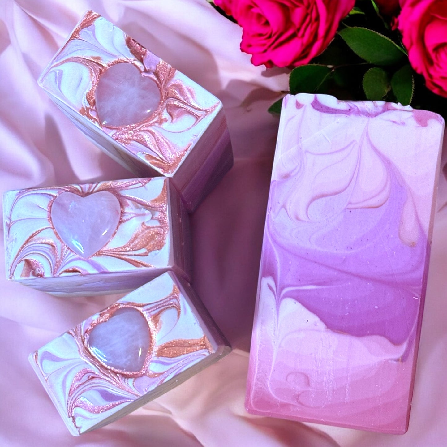 A row of rectangular bars of soap with an ombre pink to purple color gradient, displayed with a pink draped background and red roses in the background.