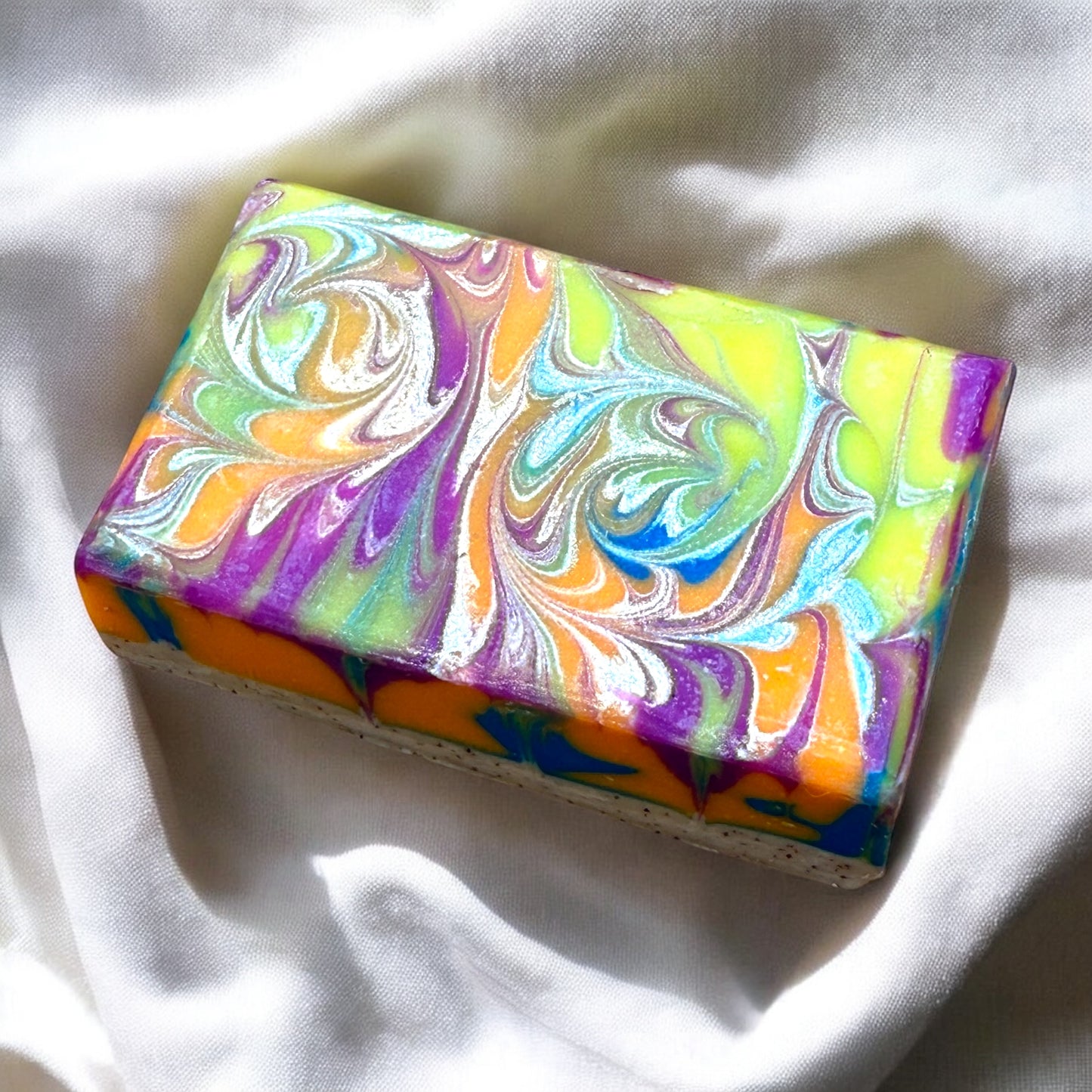 A bar of artisan soap with a multicolor swirl design, predominantly in shades of purple and yellow, placed on a white cloth surface.