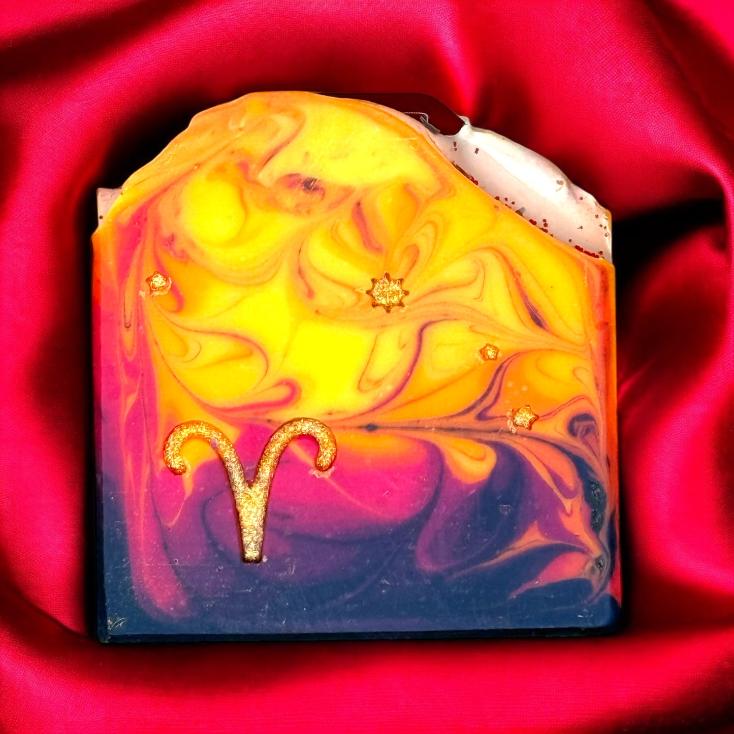 A bar of artisan soap with a swirling yellow and purple design, featuring a stamped Aries symbol and a red jasper crystal on top.