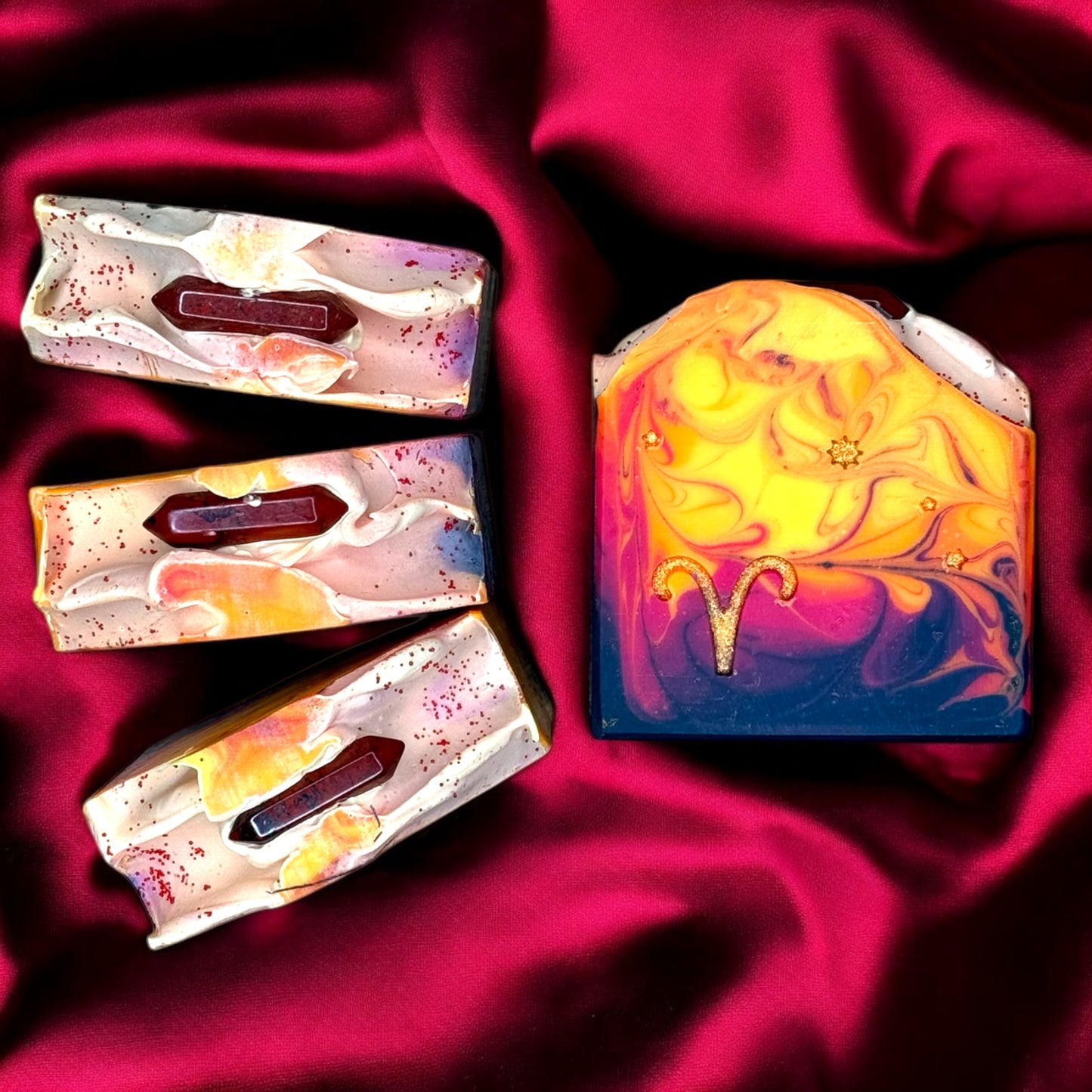 A bar of artisan soap with a swirling yellow and purple design, featuring a stamped Aries symbol and a red jasper crystal on top.