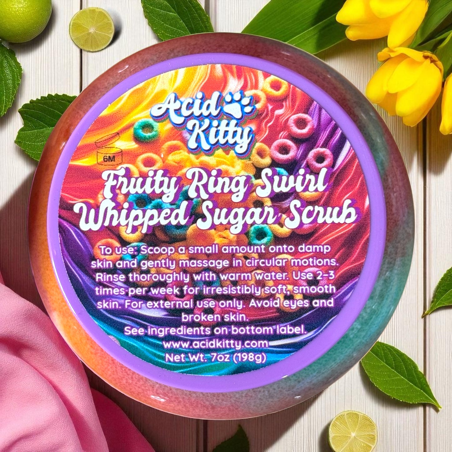 Container of Acid Kitty Fruity Ring Swirl Whipped Sugar Scrub with colorful design on a wooden surface with flowers and limes.