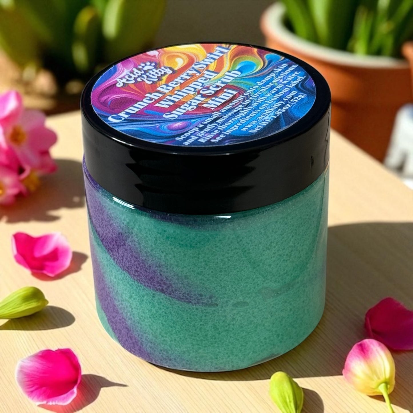 A mini jar of Crunch Berry sugar scrub on a light wooden surface surrounded by pink flower petals.