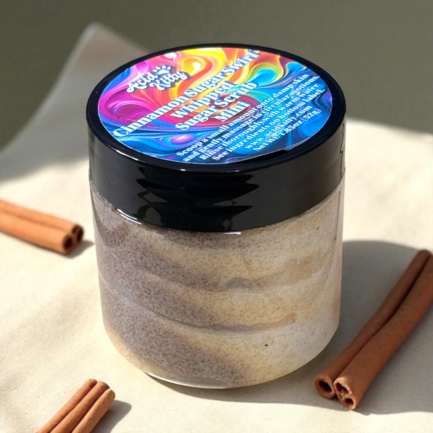 A mini jar of Cinnamon Sugar scented sugar scrub in a clear plastic jar surrounded by cinnamon sticks.