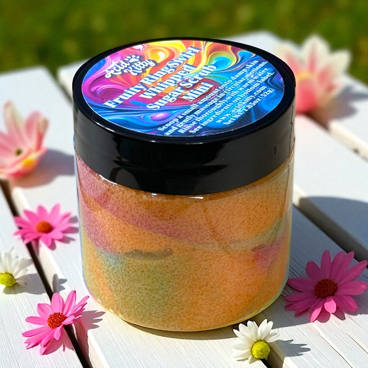 A mini jar of whipped sugar scrub with a colorful label on a wooden surface surrounded by little pink and white flowers.