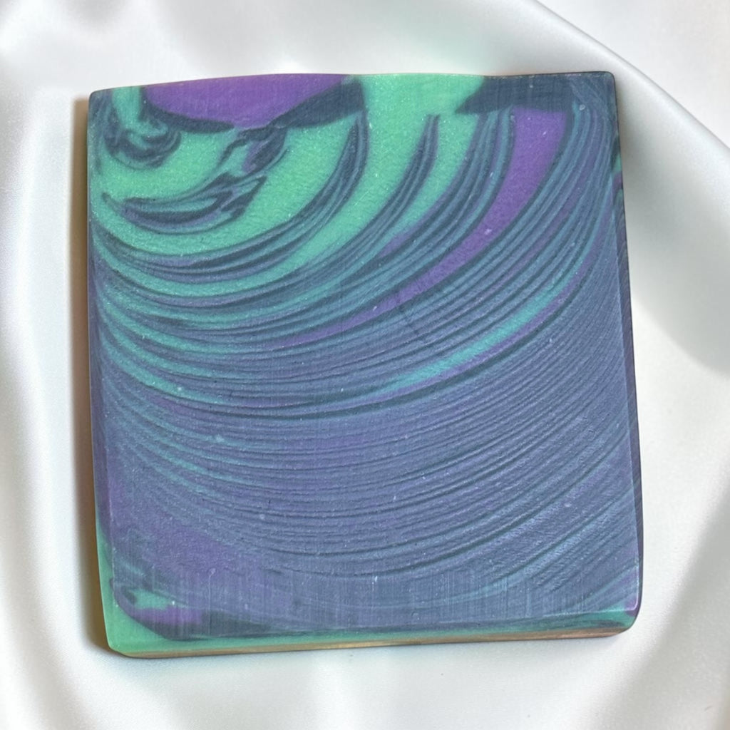 A square handmade soap bar with a marbled swirl pattern of purple, teal green, and dark blue, resting on a white satin fabric background.