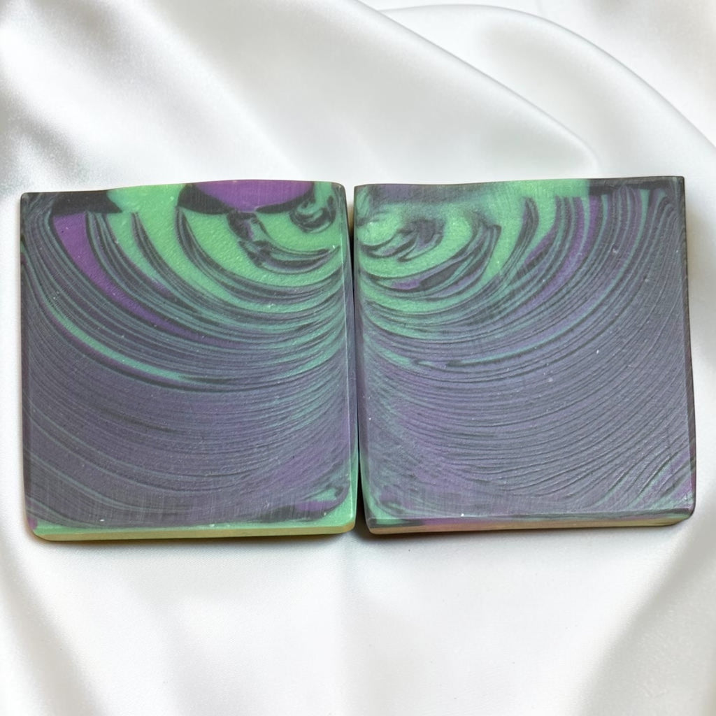 Two square bars of soap with a marbled swirl pattern of green, purple, and gray, displayed side-by-side on a white satin fabric background.