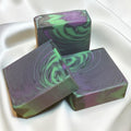 Three rectangular handmade soap bars with a marbled swirl pattern of purple, green, and dark gray, arranged on a white satin fabric background.