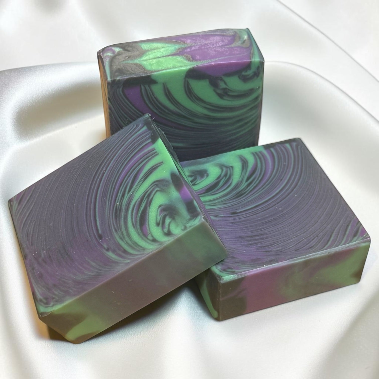 Three rectangular handmade soap bars with a marbled swirl pattern of purple, green, and dark gray, arranged on a white satin fabric background.