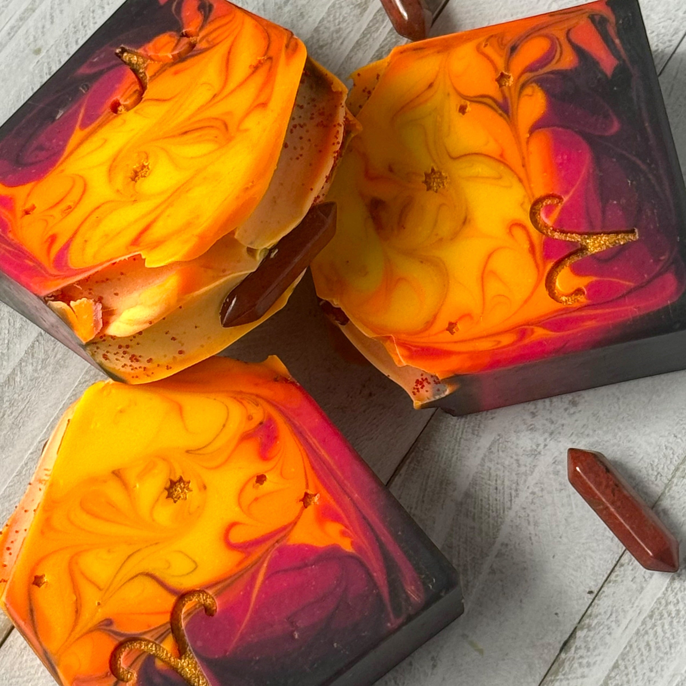 Three decorative rectangular soaps with marbled orange, yellow, red, and purple designs, arranged on a light gray wood surface with a small red crystal stake nearby.