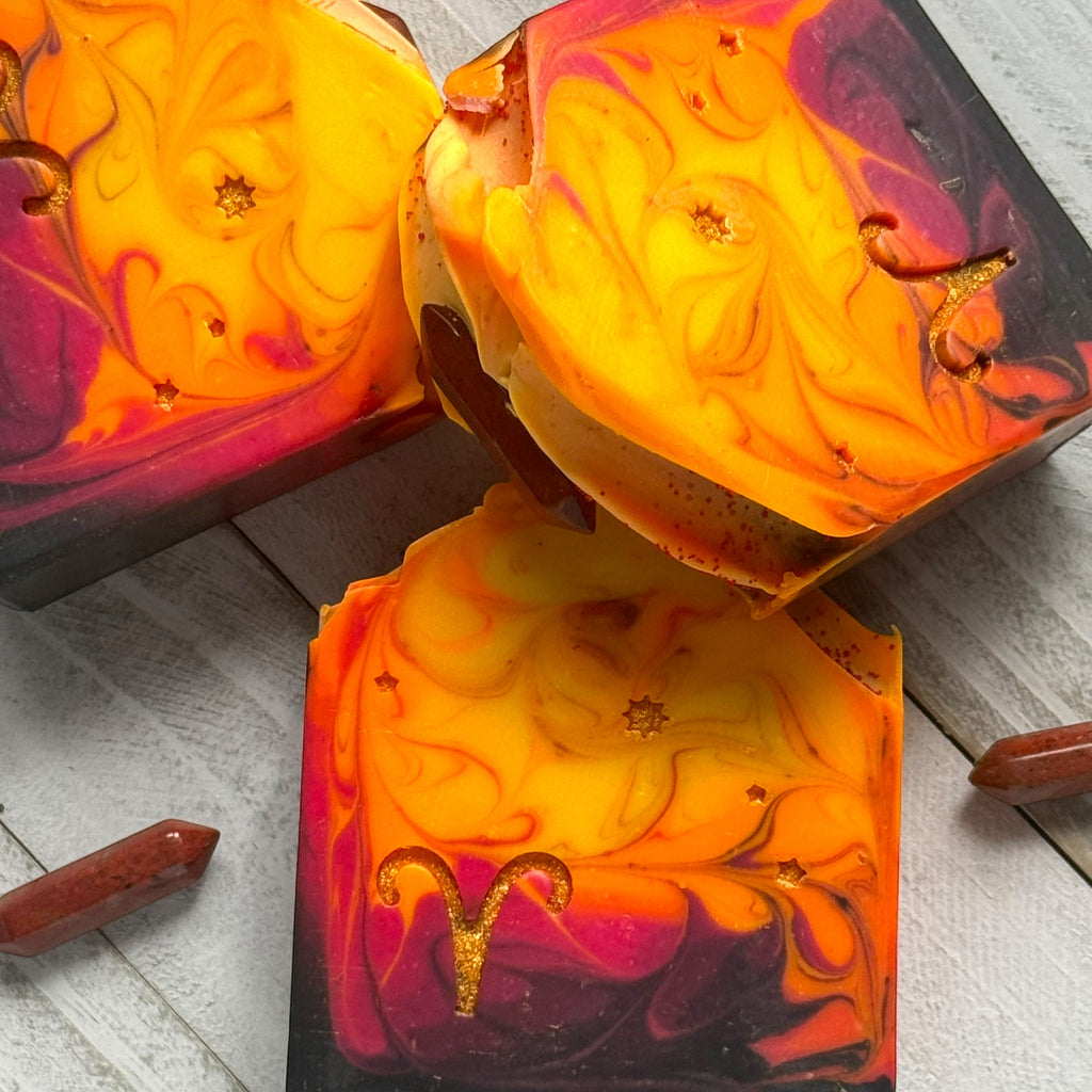 Three marbled artisanal soaps with a gold Aries zodiac symbol, featuring vibrant orange, yellow, red, and purple swirls, arranged on a light gray wood background with red crystal points.