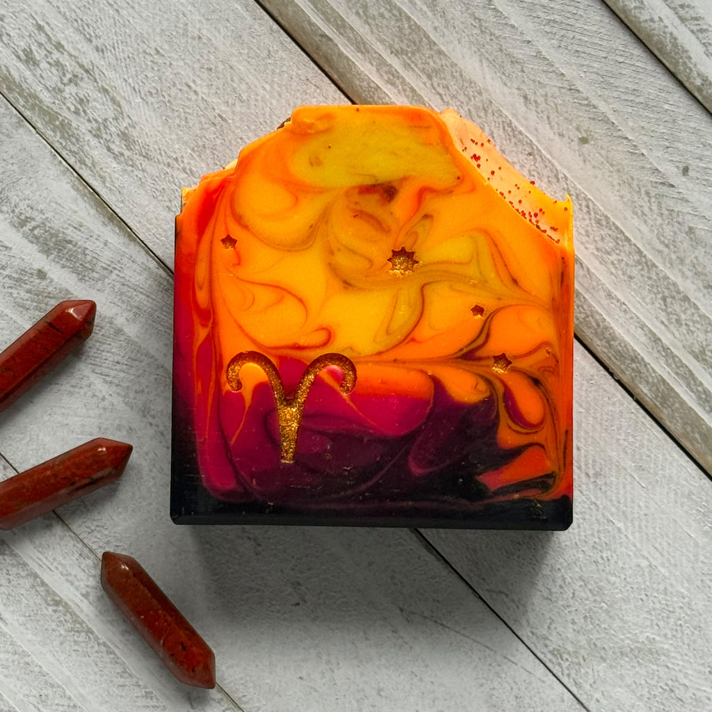 A rectangular handmade soap bar with a marbled design of orange, yellow, red, and purple, featuring a gold Aries zodiac symbol and small gold glitter flecks. The soap is displayed on a light gray wooden plank surface next to three red crystal points.
