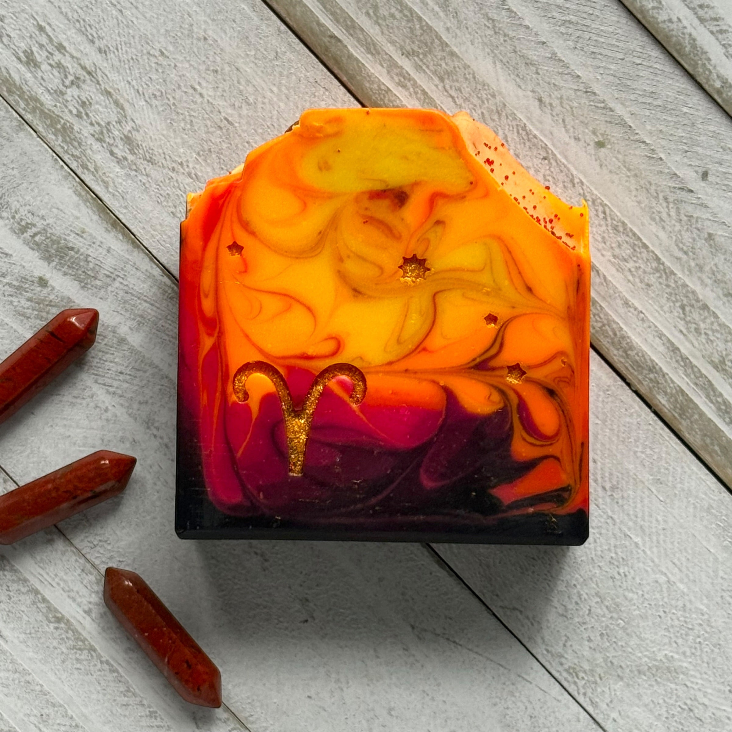 A rectangular handmade soap bar with a marbled design of orange, yellow, red, and purple, featuring a gold Aries zodiac symbol and small gold glitter flecks. The soap is displayed on a light gray wooden plank surface next to three red crystal points.