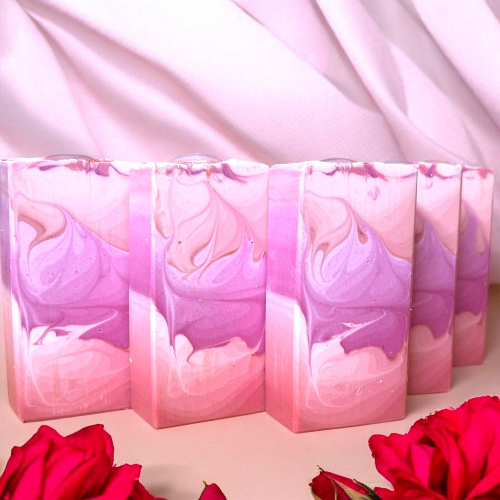 A row of rectangular bars of soap with an ombre pink to purple color gradient, displayed with a pink draped background and red roses in the foreground.