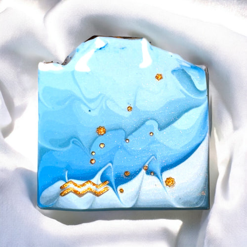 A bar of soap with a liquid blue design featuring gold stars and the Aquarius symbol stamped on the front, with a blue crystal on top.