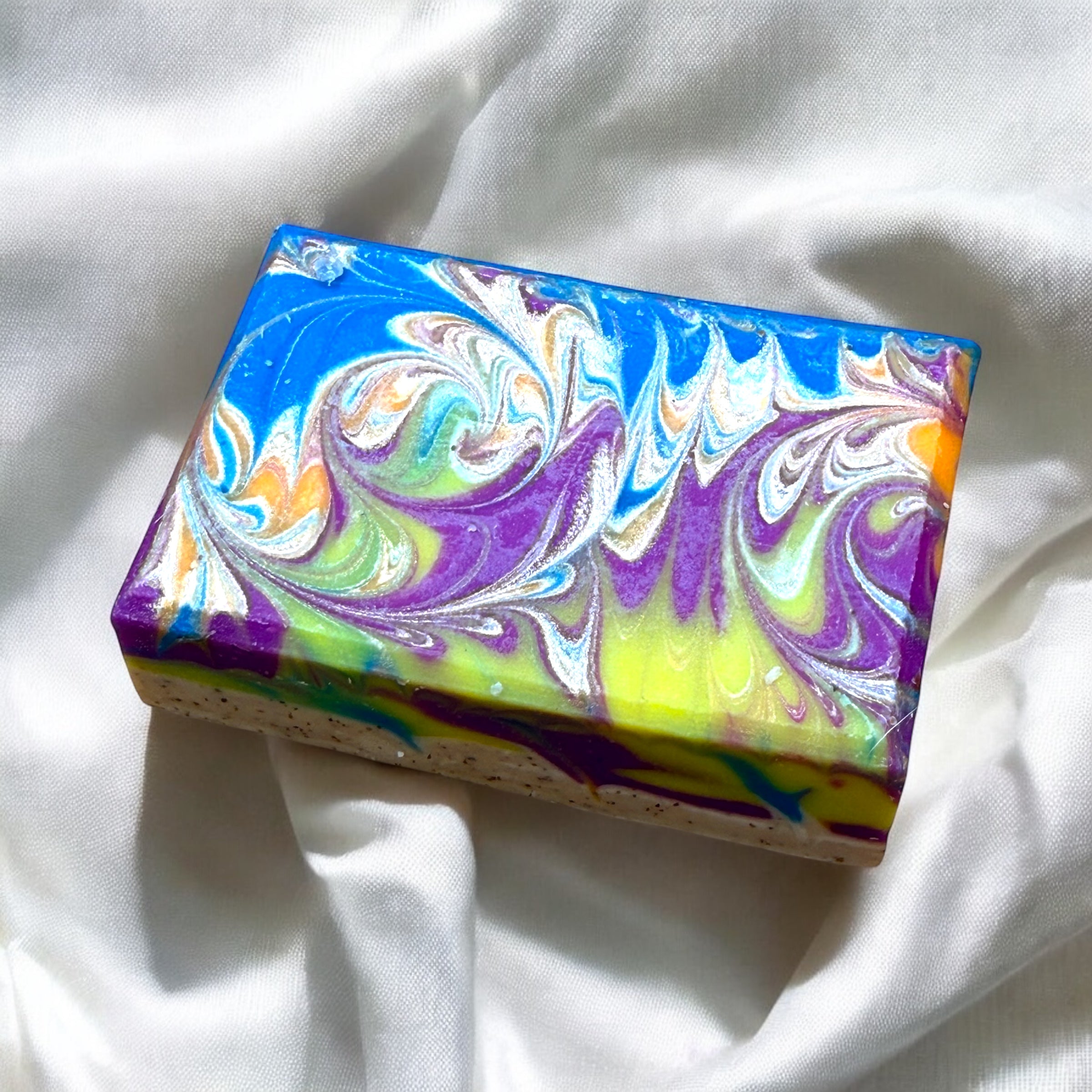 A bar of artisan soap with a multicolor swirl design, predominantly in shades of blue, purple, and yellow, placed on a marble surface.