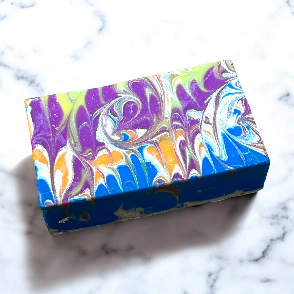A bar of artisan soap with a multicolor swirl design, predominantly in shades of blue, purple, and yellow, placed on a marble surface.