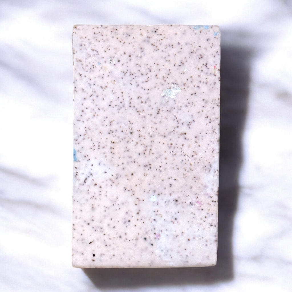 One bar of artisan soap displaying the back, which is white with flecks of walnut shell powder, displayed on a marble surface.