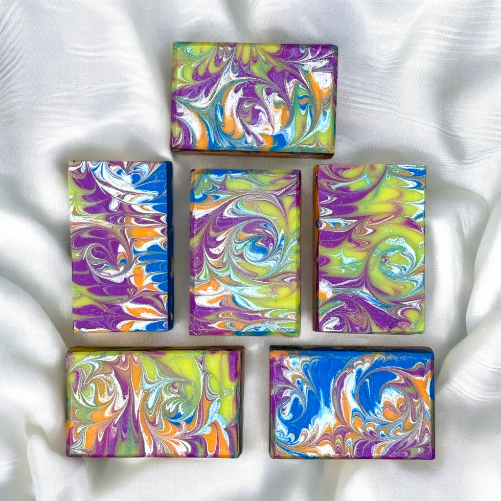 Six bars of artisan soap with a multicolor swirl design, predominantly in shades of blue, purple, and yellow, placed on a marble surface.