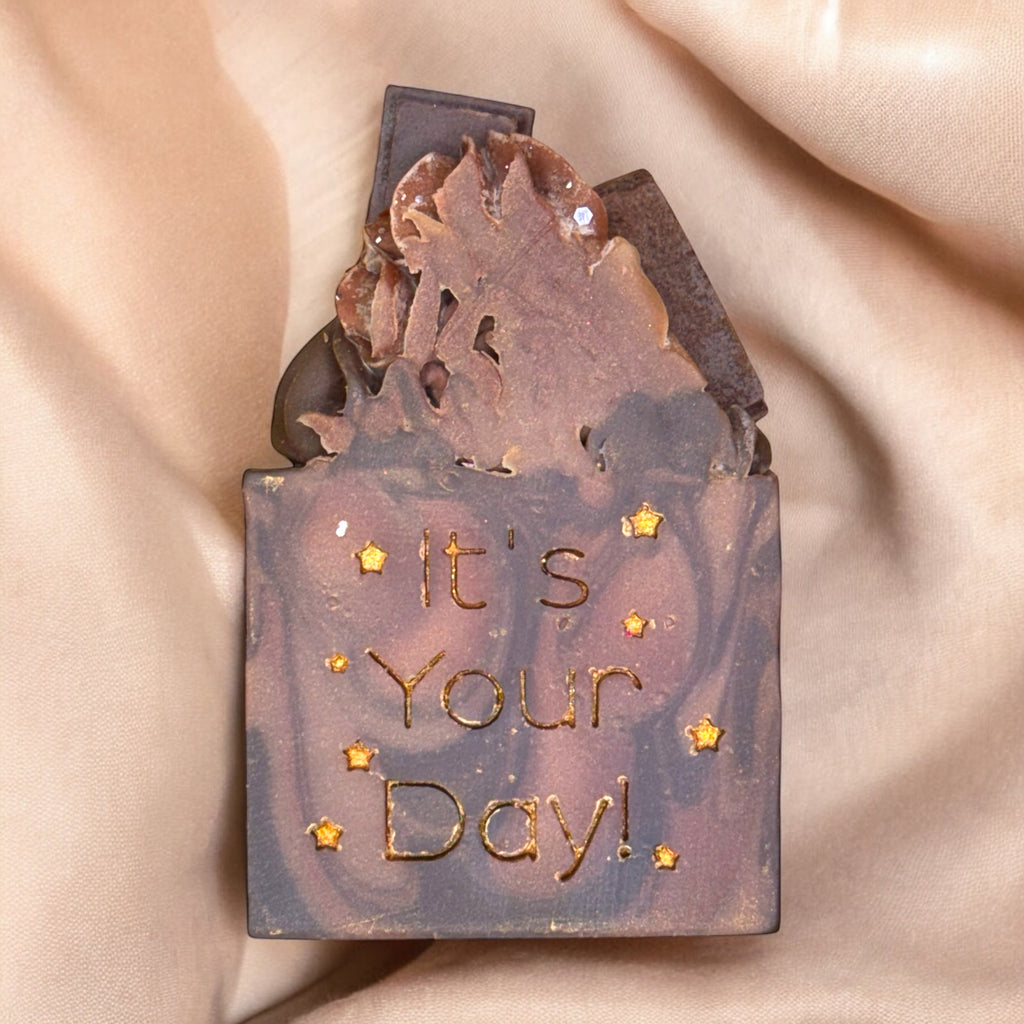 A bar of soap with a fudge brownie design, topped with mini chocolate bars, and the text 'It's Your Day!' stamped on the surface.