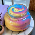 Jar of colorful whipped sugar scrub on a marble surface with a purple towel and plant in the background.