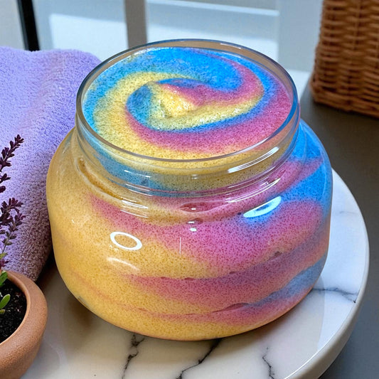 Jar of colorful whipped sugar scrub on a marble surface with a purple towel and plant in the background.
