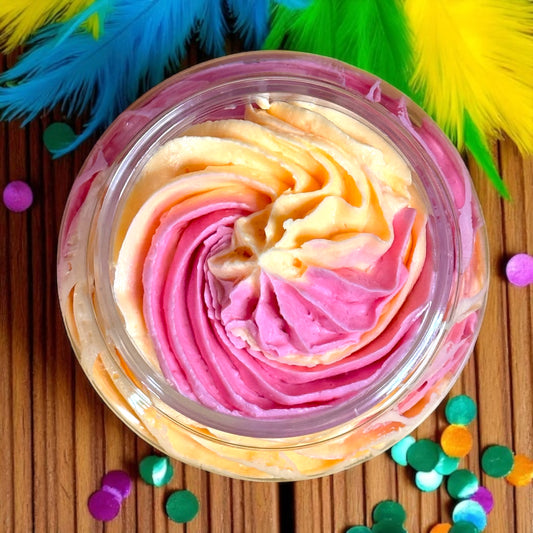 Colorful swirled whipped soap in a transparent container on a wooden surface with colorful feathers and confetti.