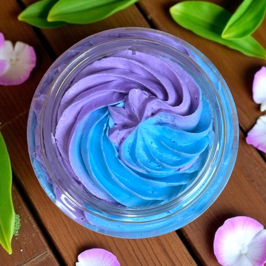 Swirl of blue and purple creamy whipped soap in a clear container on a wooden surface with green leaves and pink flowers.