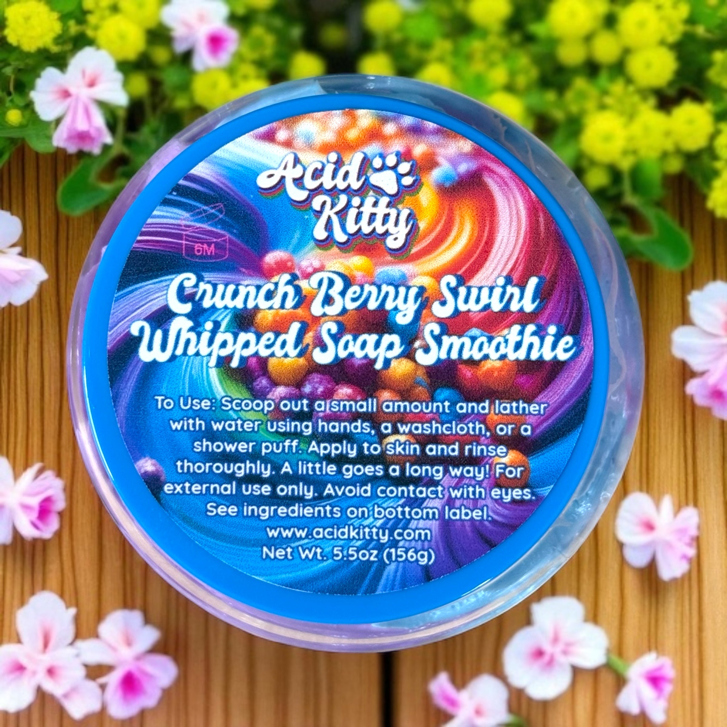 Container of Acid Kitty Crunch Berry Swirl Whipped Soap Smoothie with colorful label on a wooden surface with flowers.