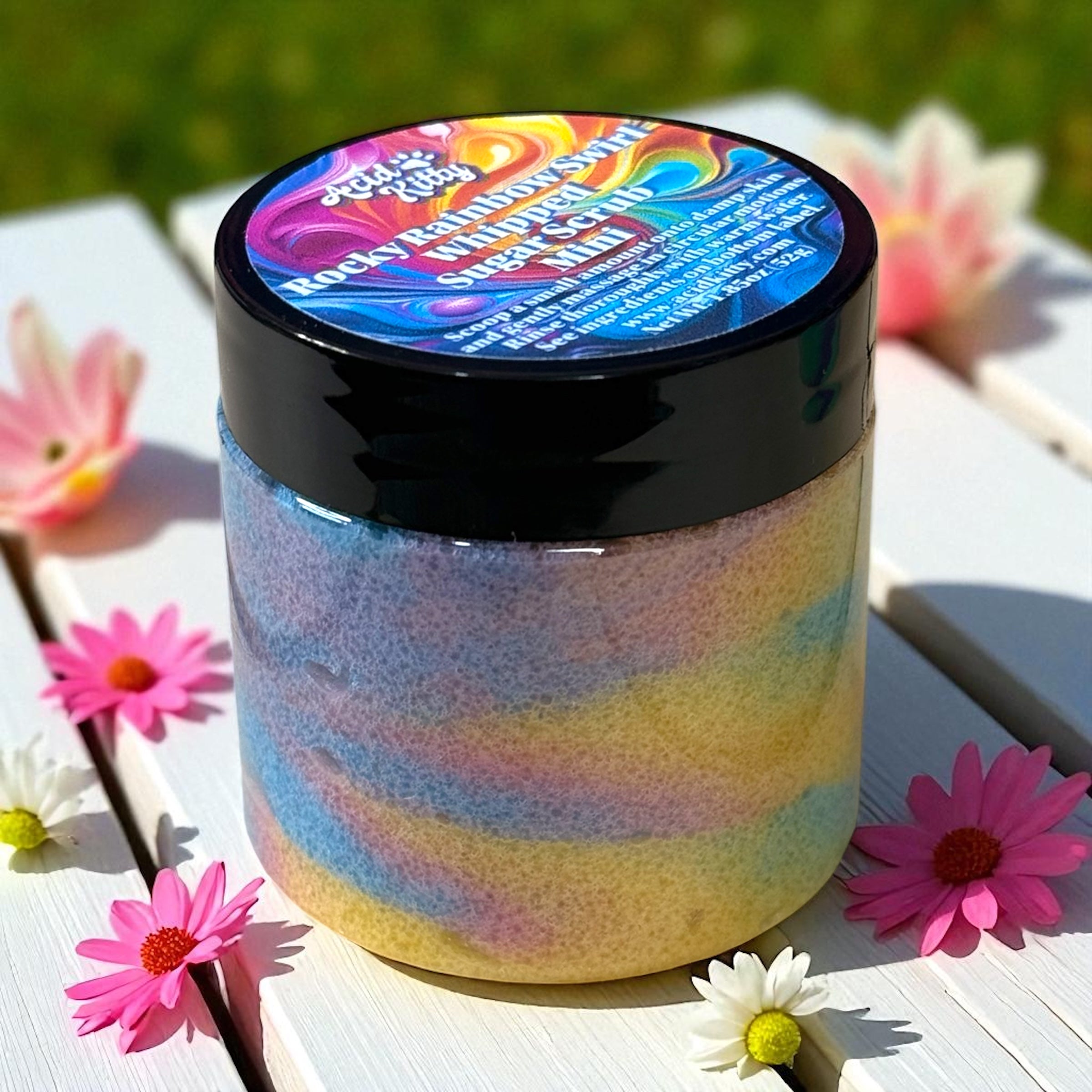 A mini jar of colorful sugar scrub on a wooden surface surrounded by little flowers. 